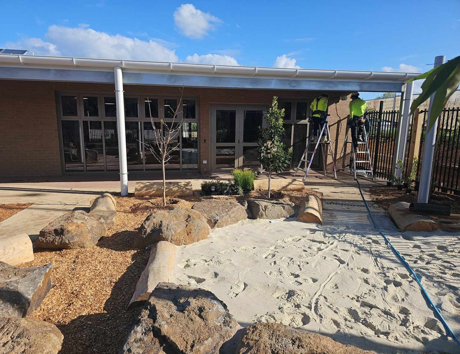 Newly landscaped courtyard of Biyala Primary School with tanbark, large stone and logs surround a sandpit. Two tradespeople are on ladders in the background.