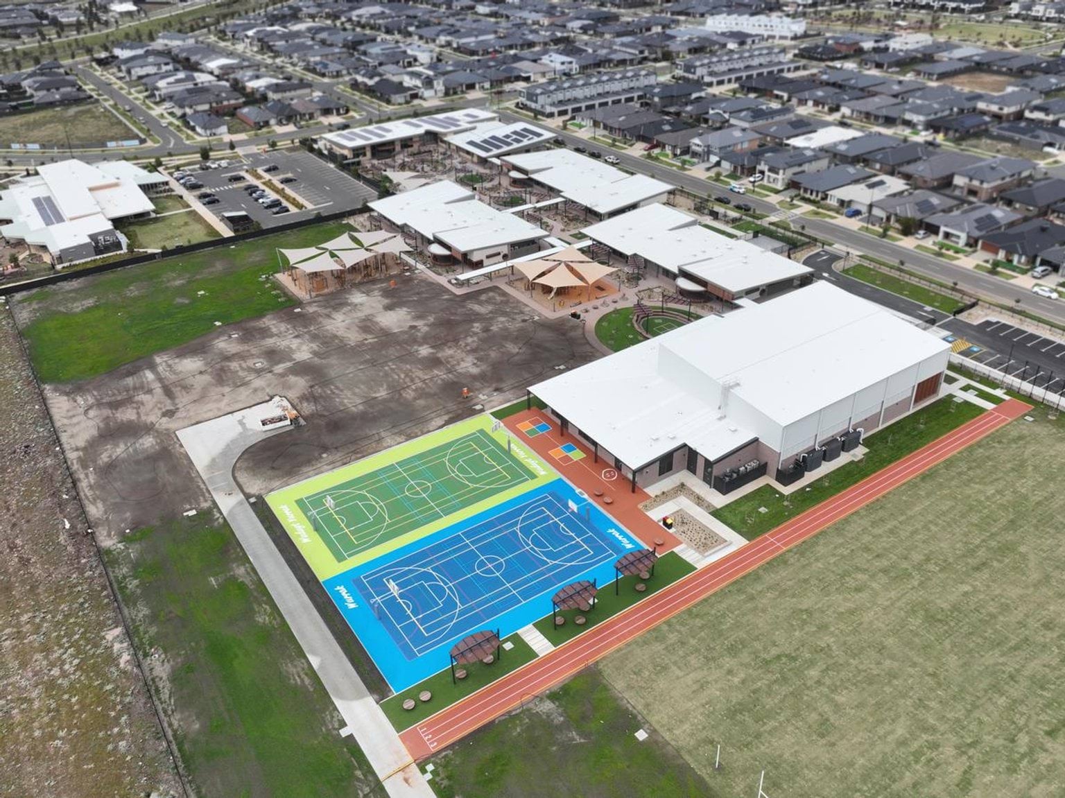 An aerial photograph showing the Mindalk Primary School construction. There is a green and blue hard court next to a building. There are other buildings that look nearly finished. The rest of the site is grass and dirt. 