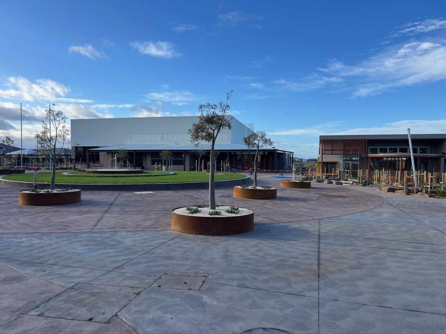 A photo of Wiyal Primary School's construction progress. There are 2 buildings in the background that look almost finished. In the foreground there is concrete pathways and planter boxes with young trees spaced throughout the pathway.