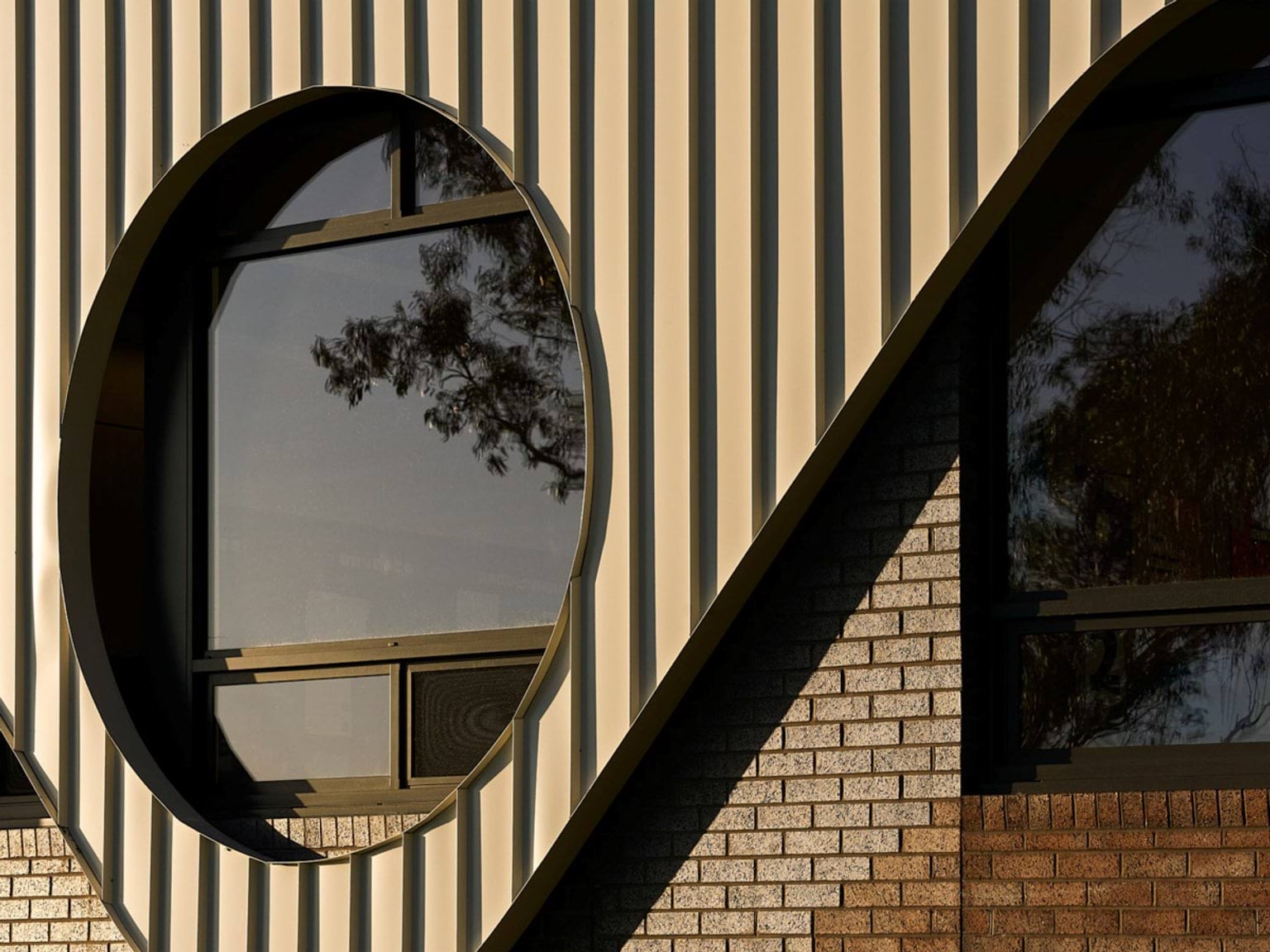 Detail of circular window framed by ribbed metal cladding and brick wall, reflecting surrounding trees