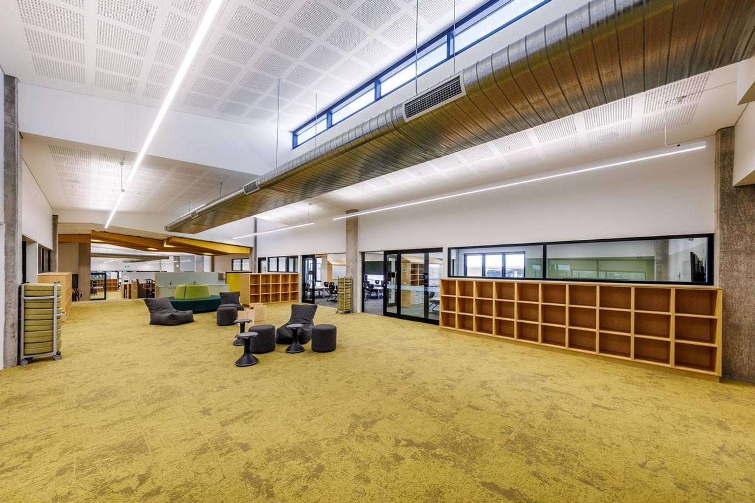 Inside Kerribana Primary School. The room's carpet is mustard yellow. There are cubbyhole shelves along the walls and stools and chairs in the middle of the room to sit on.