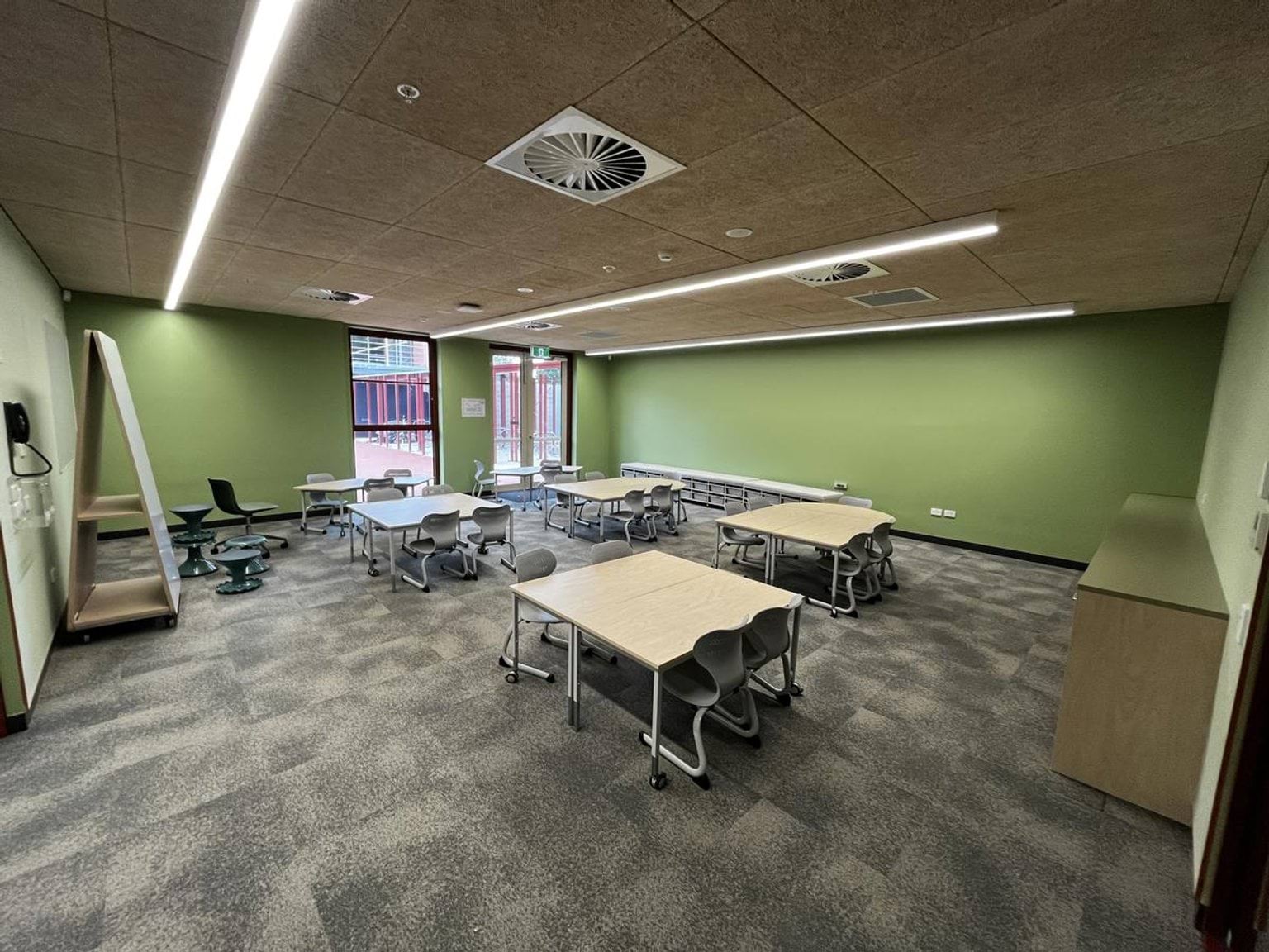A classroom with green walls and flexible furniture layout, including tables and chairs on grey carpet. Glass doors open to an outdoor area.