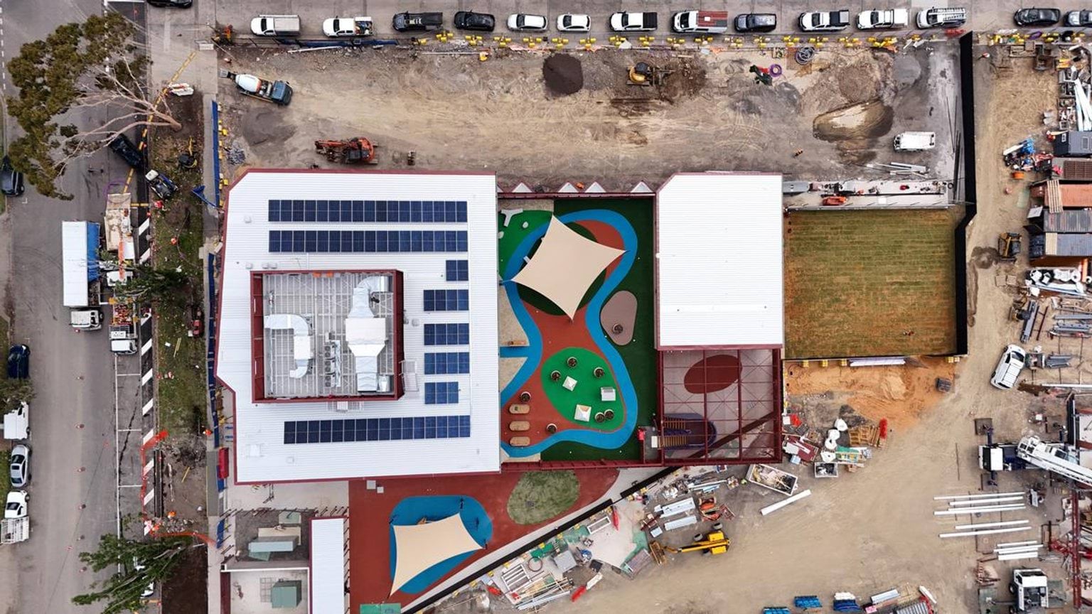 An overhead view of the entire school site with multiple buildings, colorful play areas, and construction materials spread across the grounds.