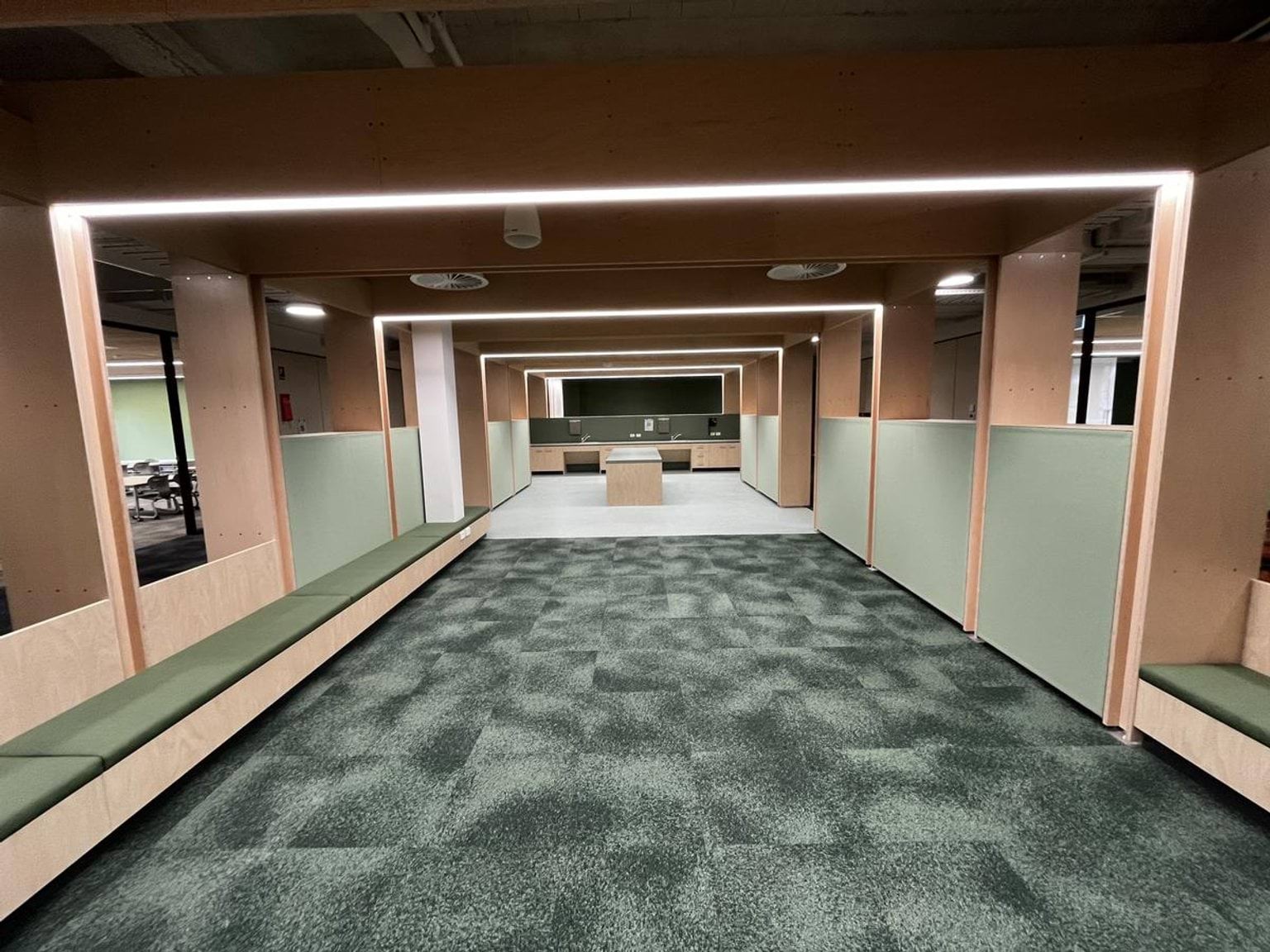 A wide interior space with green carpet, timber seating along the walls, and light panels framing the ceiling.