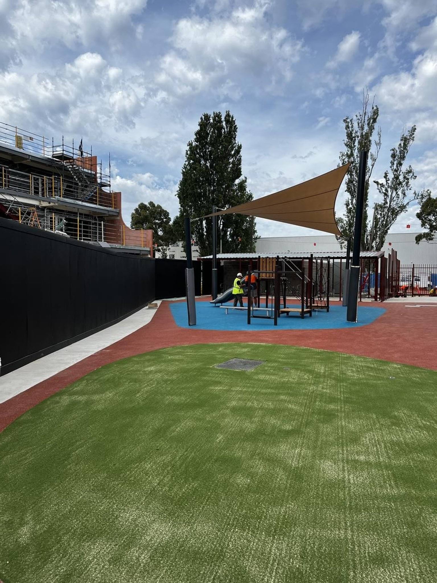 An outdoor play area with bright blue and red rubber surfacing, green artificial turf, and shade sails. Construction scaffolding is visible in the background.